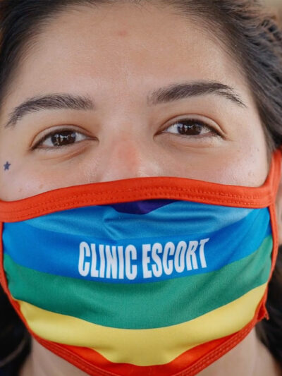 A close-up of Mariceli Alegria who's wearing a rainbow facemask with the printed words "Clinic Escort".