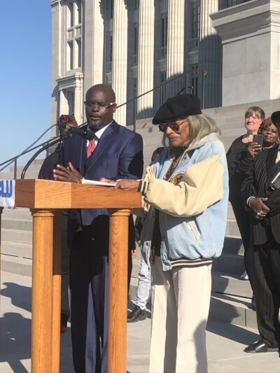 A Black man in a blue suit and a black women in a coat stand at a podium in front of a diverse crowd on the steps of the Missouri capitol buildling