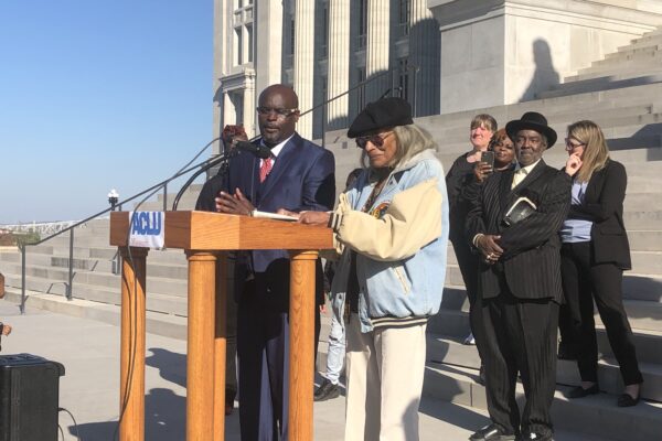 A Black man in a blue suit and a black women in a coat stand at a podium in front of a diverse crowd on the steps of the Missouri capitol buildling
