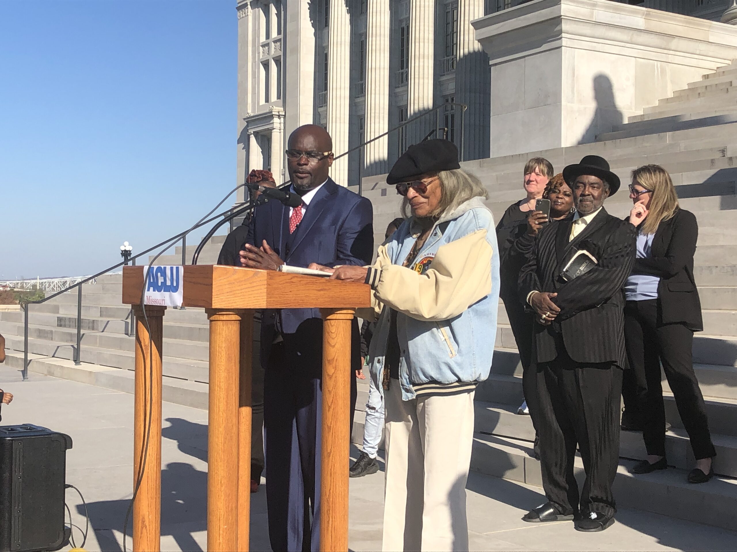 A Black man in a blue suit and a black women in a coat stand at a podium in front of a diverse crowd on the steps of the Missouri capitol buildling