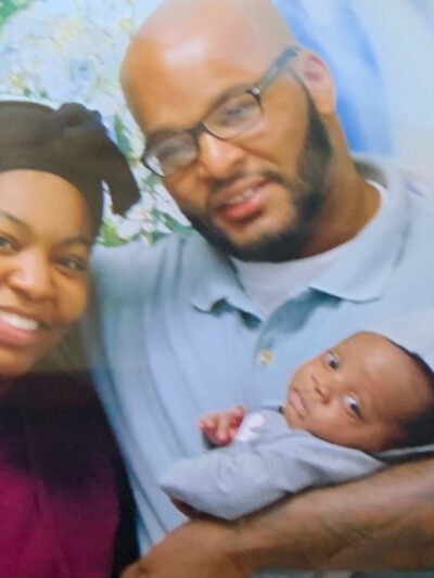 A black woman wearing a purple shirt and a black hat lovingly poses with her father while he hold her baby, his grandchild..