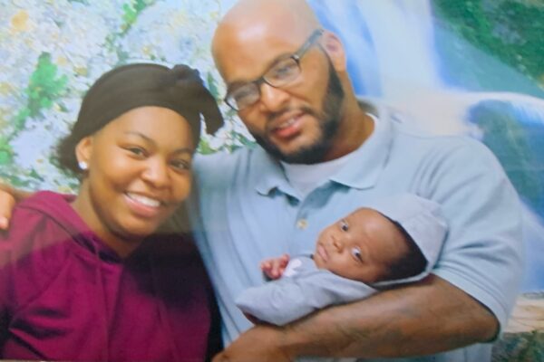A black woman wearing a purple shirt and a black hat lovingly poses with her father while he hold her baby, his grandchild..