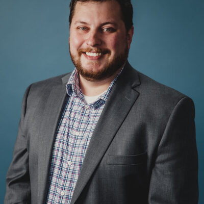 A man with short hair and a beard wearing a plaid shirt underneath a grey coat.