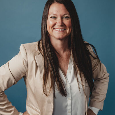 A woman with dark hair wearing a white shirt under a beige pink coat with a blue background