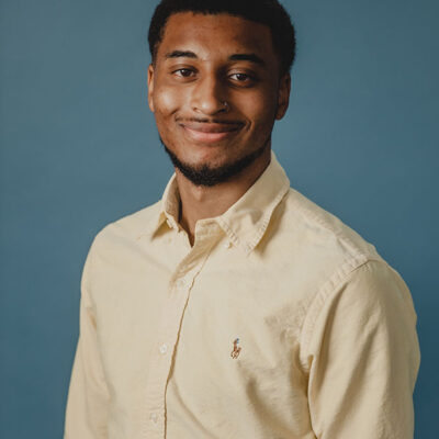 A man with dark hair and a beard wearing a cream shirt with a blue background.