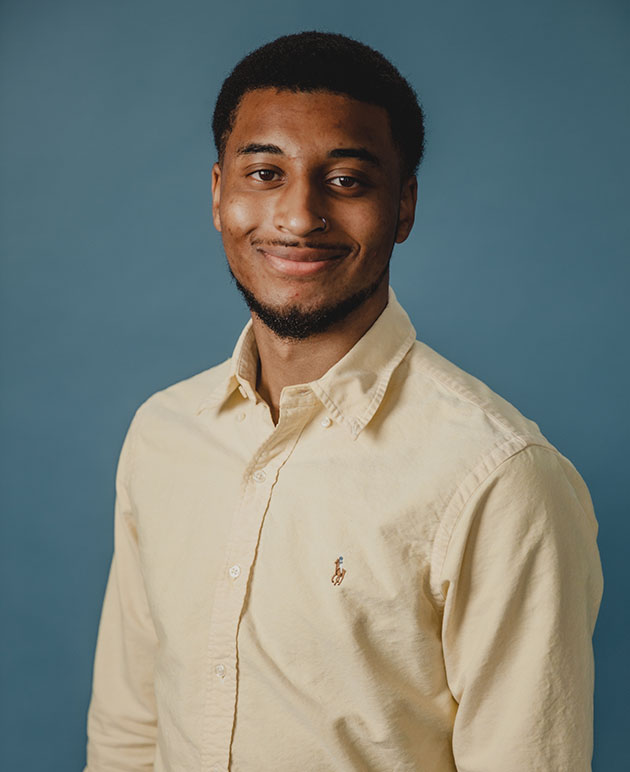 A man with dark hair and a beard wearing a cream shirt with a blue background.