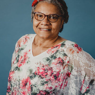 A woman with dark glasses wearing a red bow and a floral blouse with a blue background.