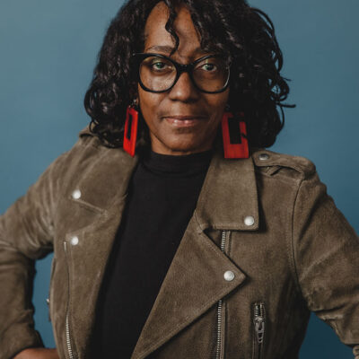 A black woman with shoulder length hair wearing a grey coat, red earrings and black framed glasses.