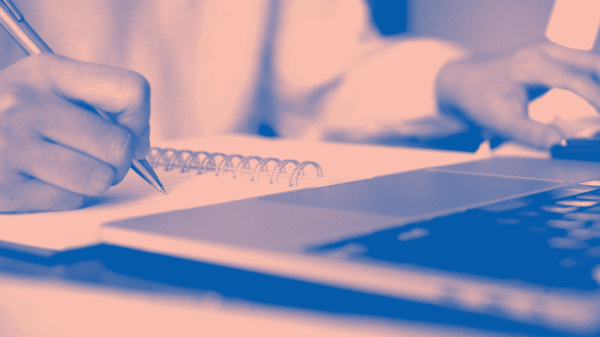 blue and light pink image, close up of a white person writing in notebook and browsing on laptop