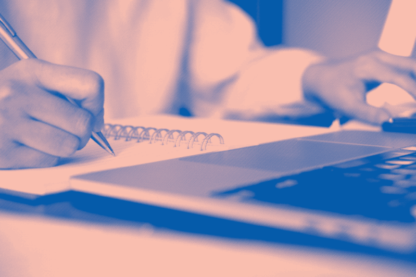 blue and light pink image, close up of a white person writing in notebook and browsing on laptop