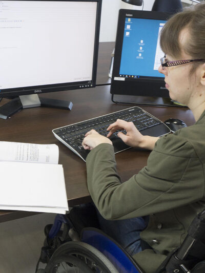 Disabled worker in a wheelchair typing at keyboard while looking at two computer screens.