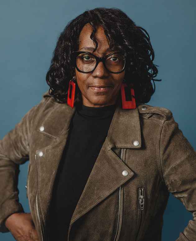 A black woman with shoulder length hair wearing a grey coat, red earrings and black framed glasses. 