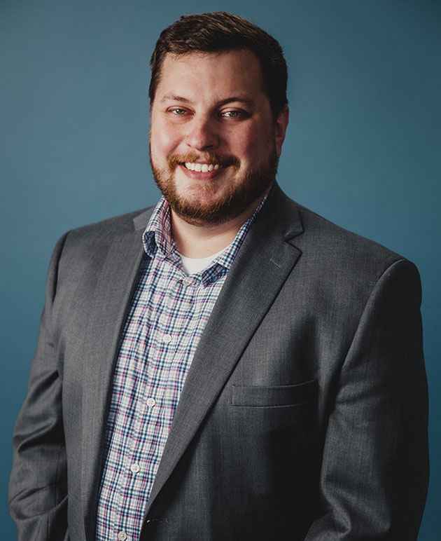 A man with short hair and a beard wearing a plaid shirt underneath a grey coat. 