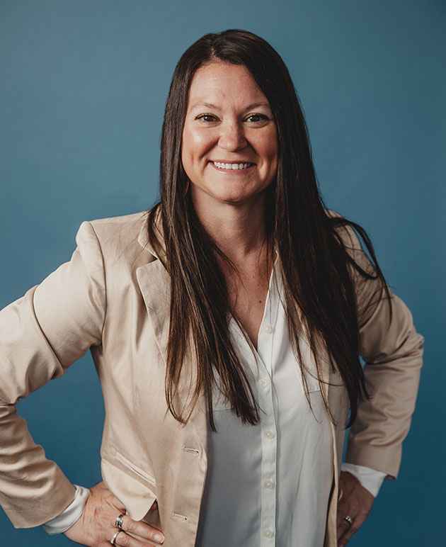 A woman with dark hair wearing a white shirt under a beige pink coat with a blue background