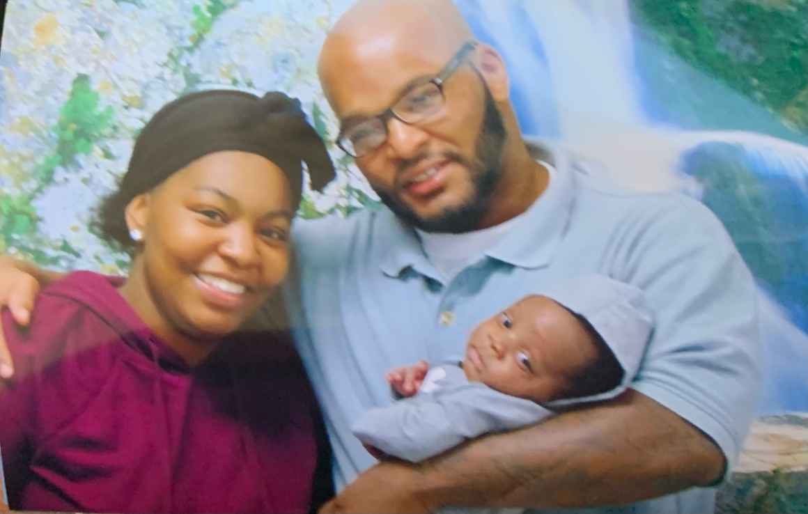 A black woman wearing a purple shirt and a black hat lovingly poses with her father while he hold her baby, his grandchild.. 