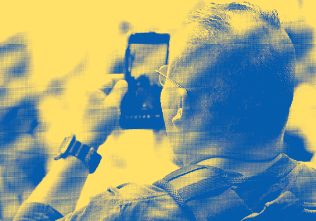 yellow and blue image, middle aged white man wearing backpack recording with his phone