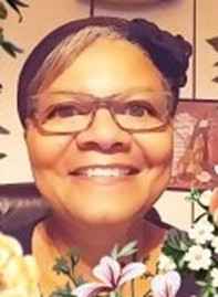 an image of a black woman wearing glasses sitting at a desk behind white and purple flowers