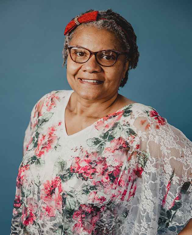 A woman with dark glasses wearing a red bow and a floral blouse with a blue background.