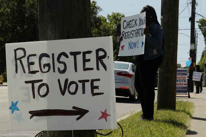 In the foreground, posted on a telephone pole reads a sign "REGISTER TO VOTE", while a woman holds a sign saying "CHECK YOUR REGISTRATION NOW!" is in the background.