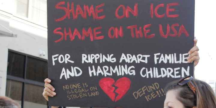 A demonstrator protests ICE and the Trump Administration policy of separating migrant families by holding up a sign (written in red ink on a black background with an illustrated broken red heart at the bottom) that reads, "SHAME ON ICE SHAME ON THE USA...