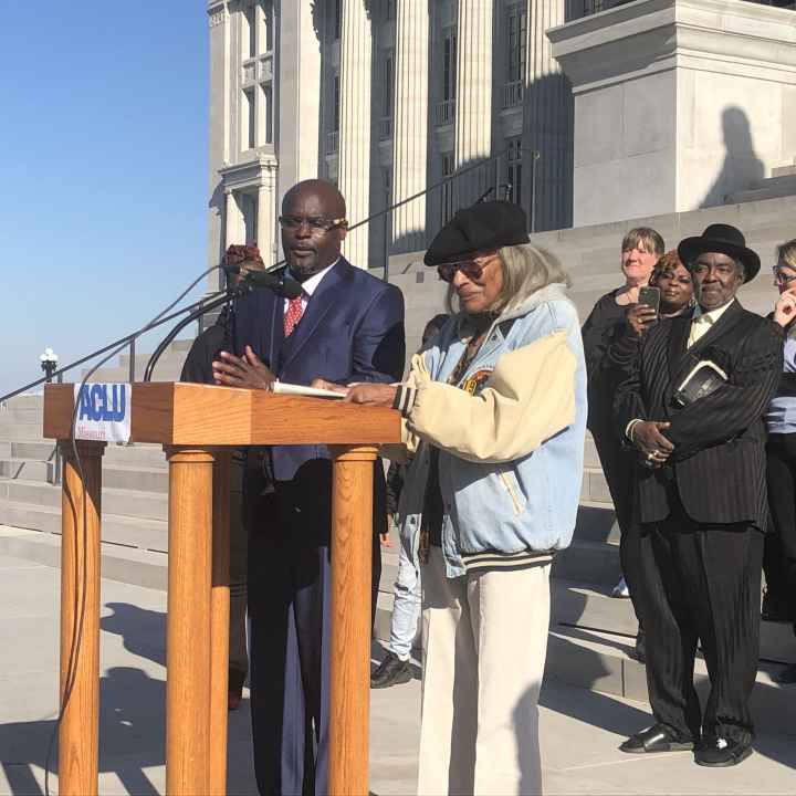 A Black man in a blue suit and a black women in a coat stand at a podium in front of a diverse crowd on the steps of the Missouri capitol buildling