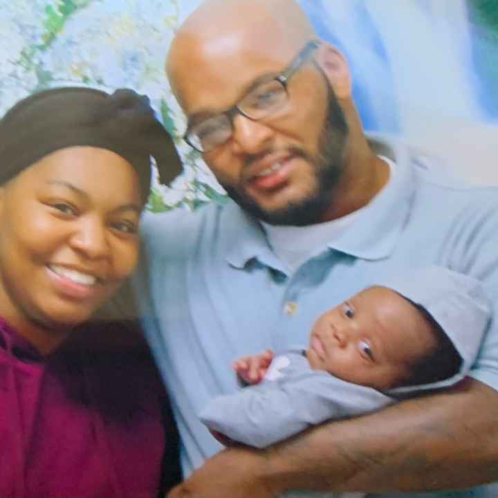 A black woman wearing a purple shirt and a black hat lovingly poses with her father while he hold her baby, his grandchild..