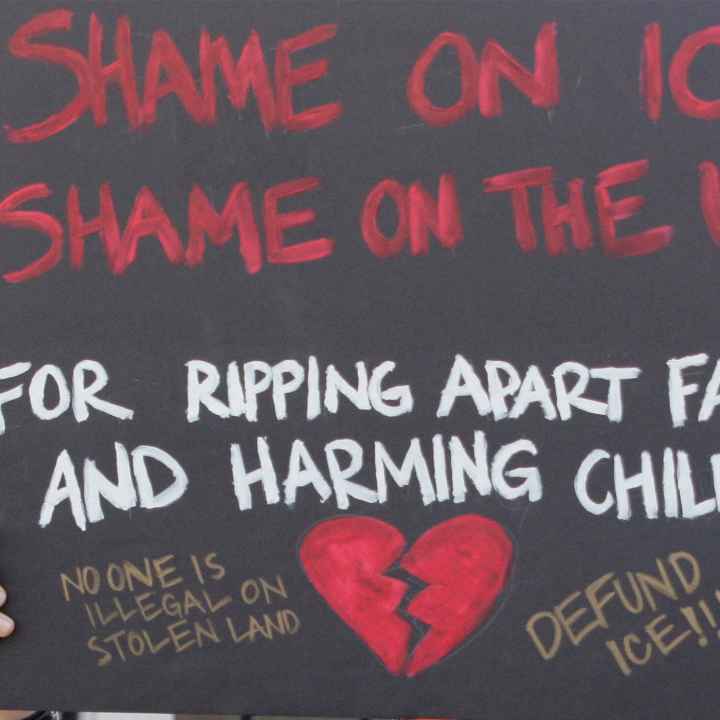 A demonstrator protests ICE and the Trump Administration policy of separating migrant families by holding up a sign (written in red ink on a black background with an illustrated broken red heart at the bottom) that reads, "SHAME ON ICE SHAME ON THE USA...